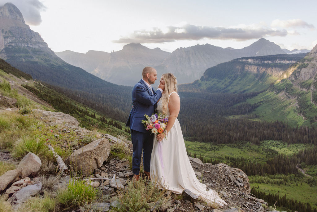 A couple stands on a rocky ledge in formal attire, with mountains and a valley in the background. The man is adjusting the woman's hair as she holds a bouquet for a wedding at glacier national park
