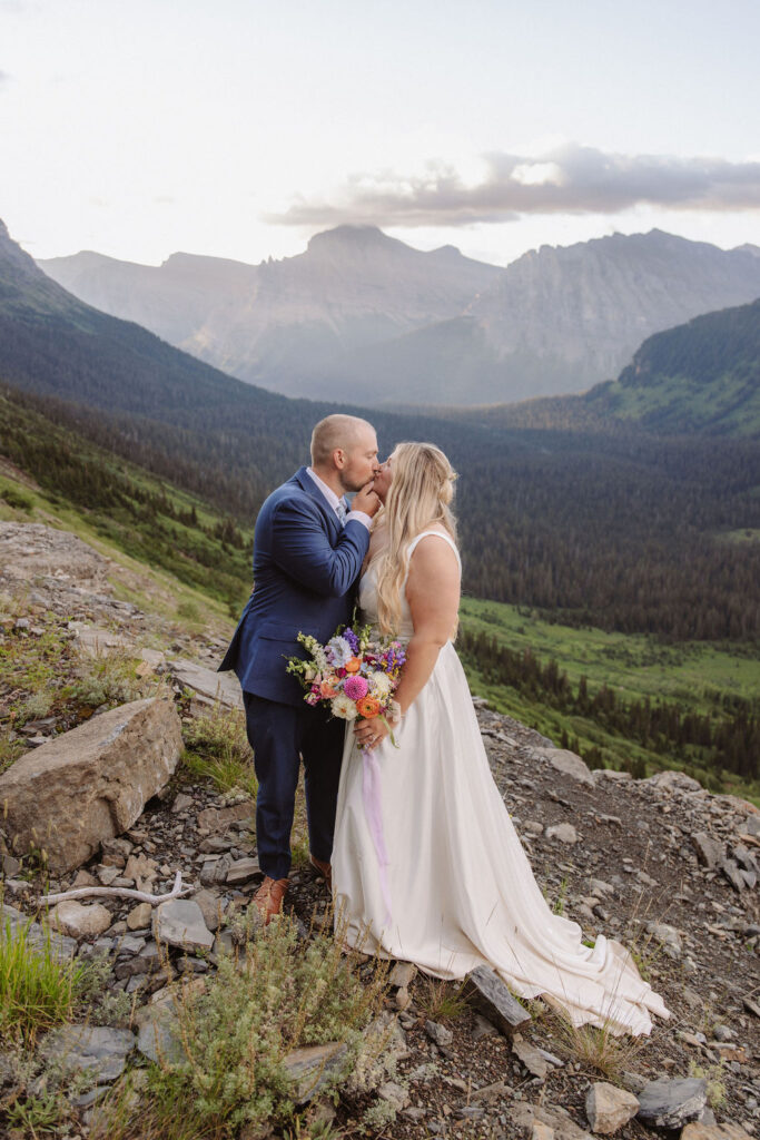 A bride and groom stand on rocky terrain overlooking a large lake and mountains, dressed in wedding attire, with trees and water in the background for a wedding at glacier national park