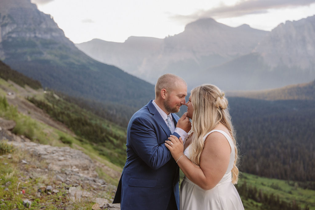 A bride and groom stand on rocky terrain overlooking a large lake and mountains, dressed in wedding attire, with trees and water in the background for a wedding at glacier national park
