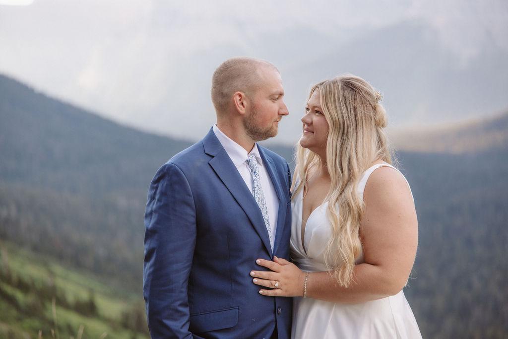 A couple dressed in formal attire stands closely together outdoors, with mountains and greenery in the background or a wedding at glacier national park