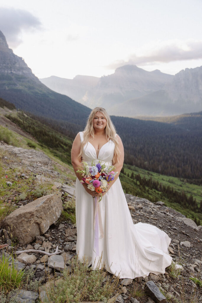 A bride and groom stand on rocky terrain overlooking a large lake and mountains, dressed in wedding attire, with trees and water in the background for a wedding at glacier national park