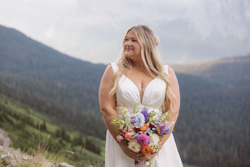A woman in a white wedding dress holds a colorful bouquet while standing outdoors with mountains and greenery in the background or a wedding at glacier national park