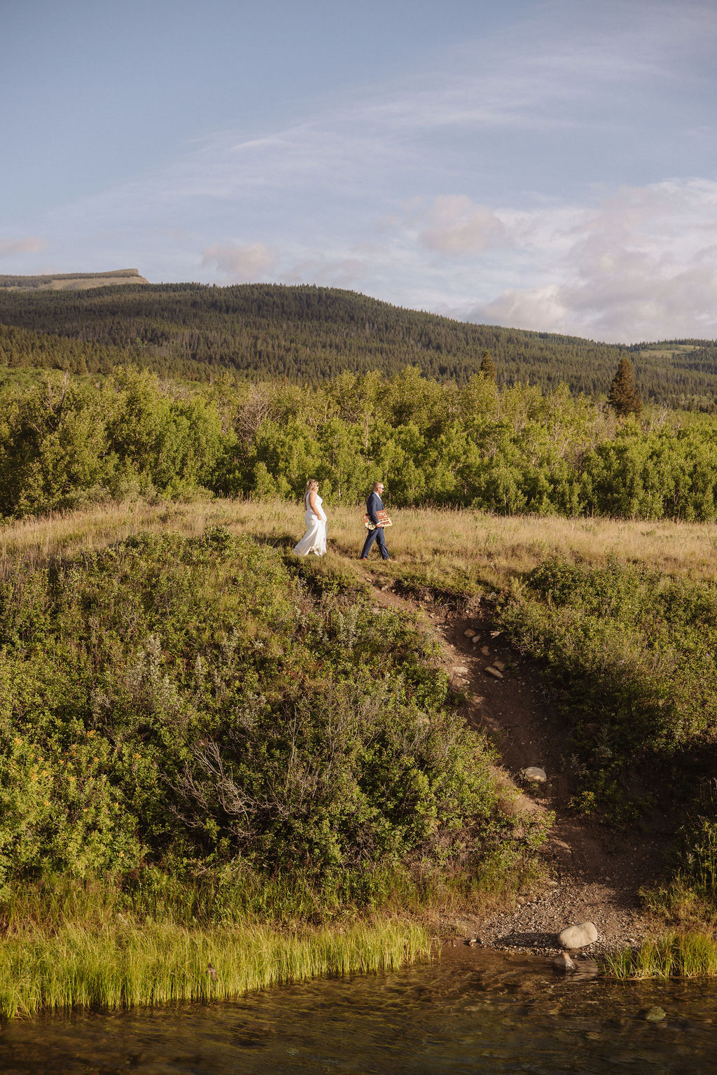 Two people walk along a grassy hillside above a body of water, with green shrubs and forested hills in the background under a partly cloudy sky or a wedding at glacier national park