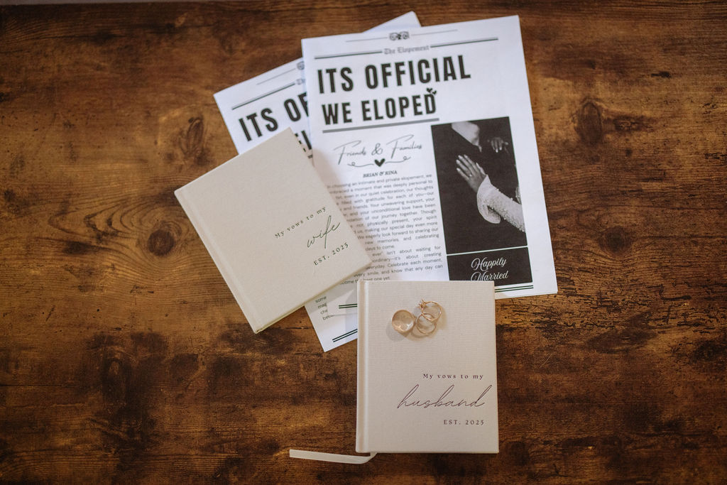 Two booklets and two newspaper-style announcements reading "It's Official We Eloped" are displayed on a wooden surface with a pair of wedding rings on one booklet.