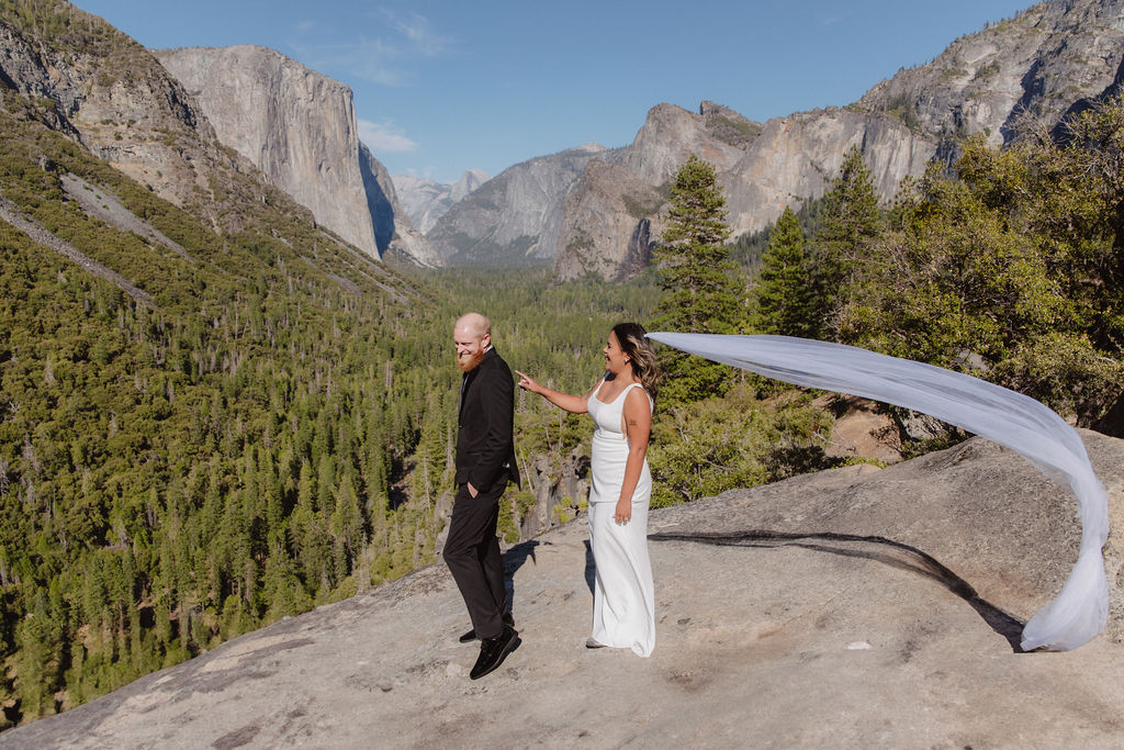 A bride and groom stand on a rocky ledge overlooking a forested valley and mountains, with the bride’s veil blowing in the wind.