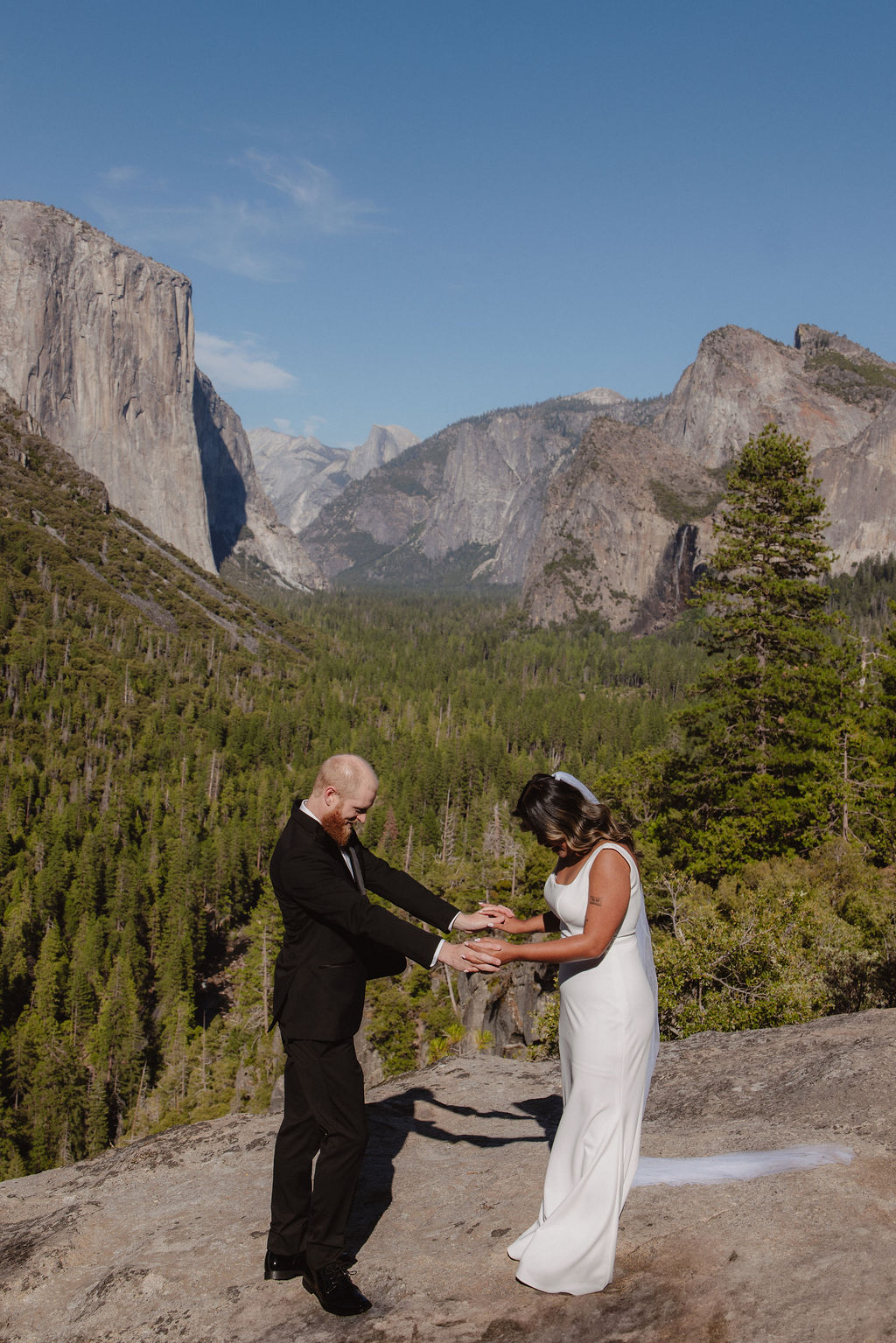 A bride and groom stand on a rocky ledge overlooking a forested valley and mountains, with the bride’s veil blowing in the wind.