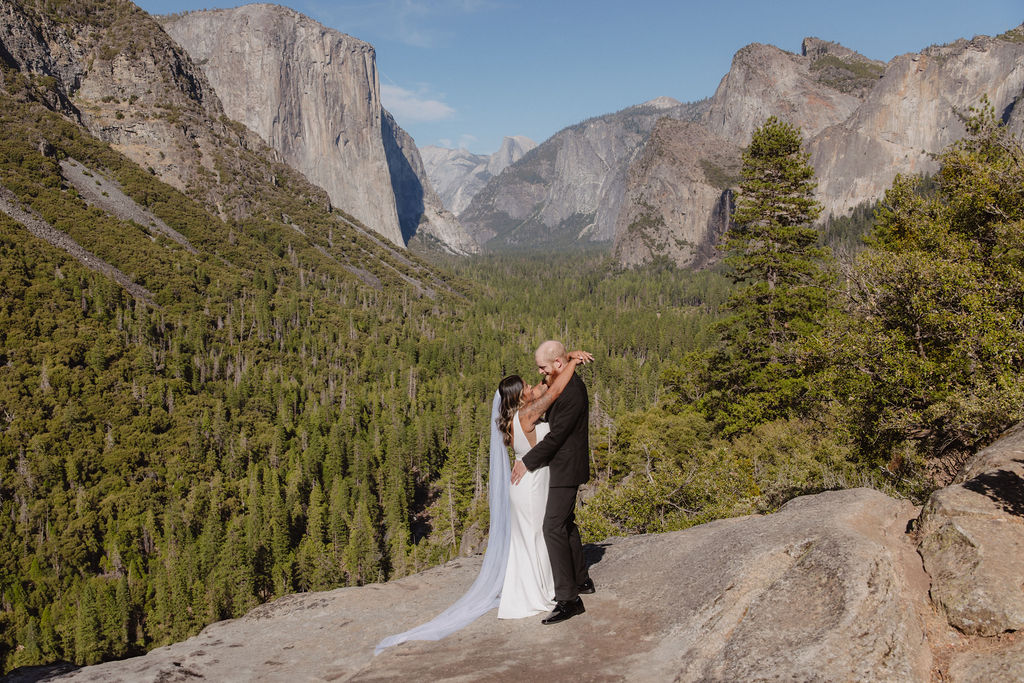 A bride and groom stand on a rocky ledge overlooking a forested valley and mountains, with the bride’s veil blowing in the wind.