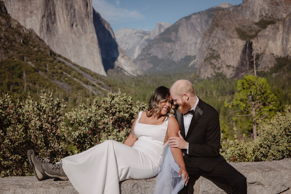 A couple dressed in wedding attire sits on a stone ledge with mountains and trees in the background under a blue sky.