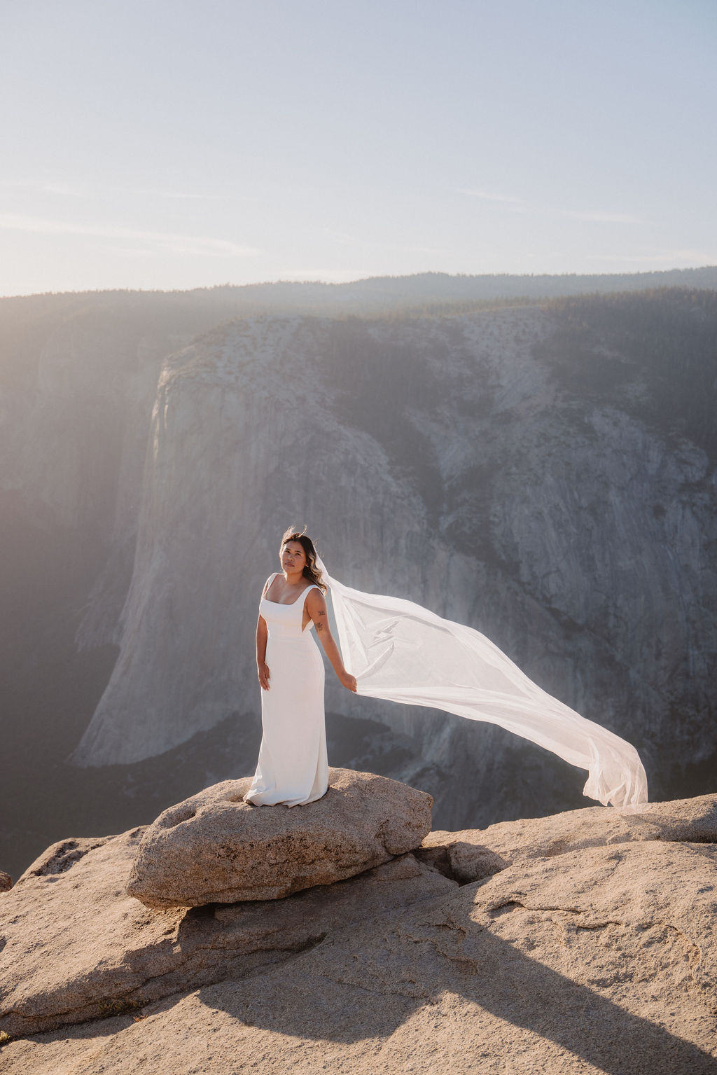 A woman in a white wedding dress stands on a rocky cliff with a long veil flowing behind her, overlooking a canyon or valley under a clear sky.