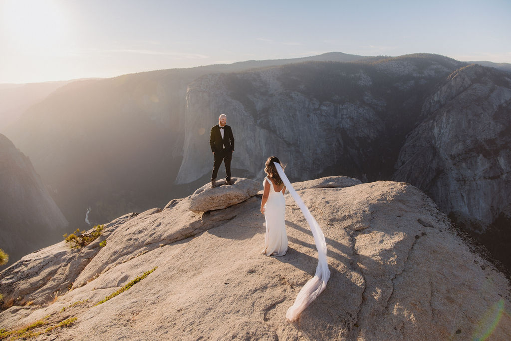 A bride and groom stand on a rocky cliff edge with mountains in the background, the bride's veil trailing behind her in the wind for a glacier point elopement