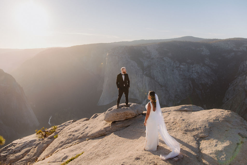 A bride and groom stand on a rocky cliff edge with mountains in the background, the bride's veil trailing behind her in the wind for a glacier point elopement