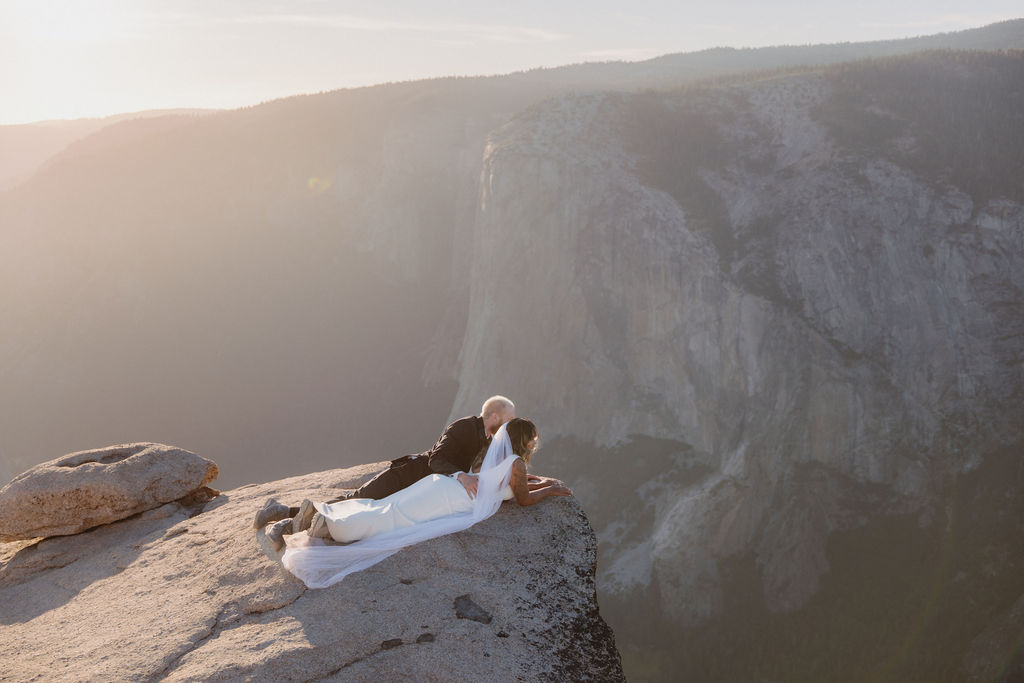 A bride and groom stand on a rocky cliff edge with mountains in the background, the bride's veil trailing behind her in the wind for a glacier point elopement