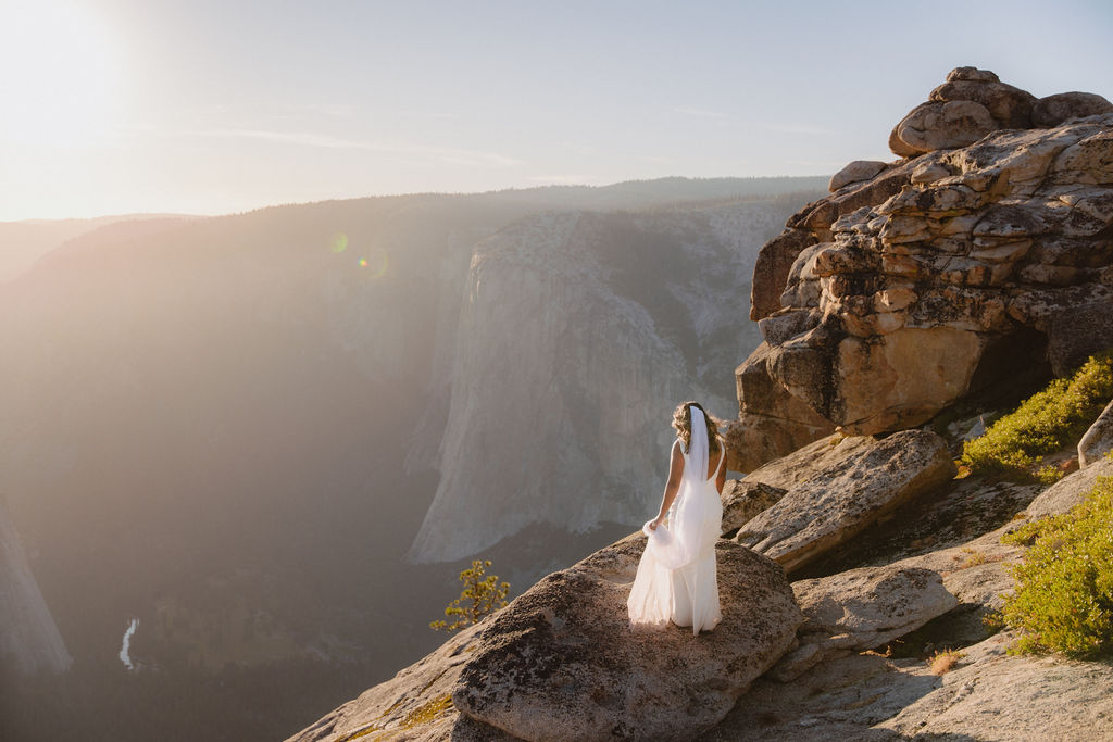 A bride and groom stand on a rocky cliff edge with mountains in the background, the bride's veil trailing behind her in the wind for a glacier point elopement