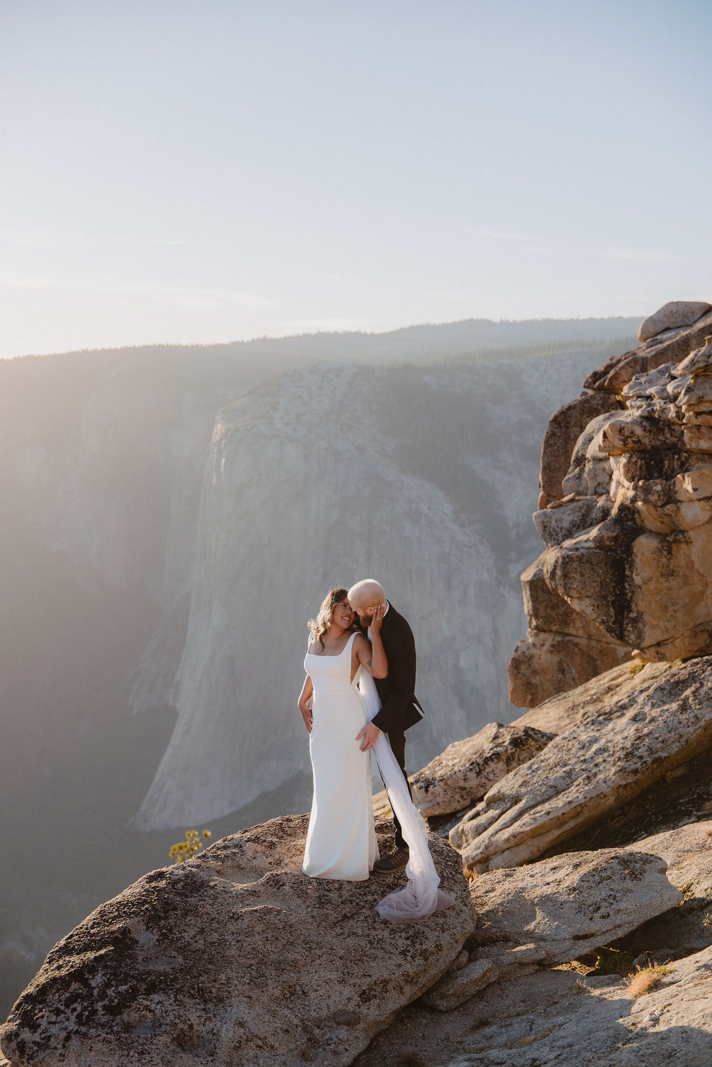 A woman in a white dress and veil stands on a rocky ledge overlooking a vast mountain landscape at sunset.