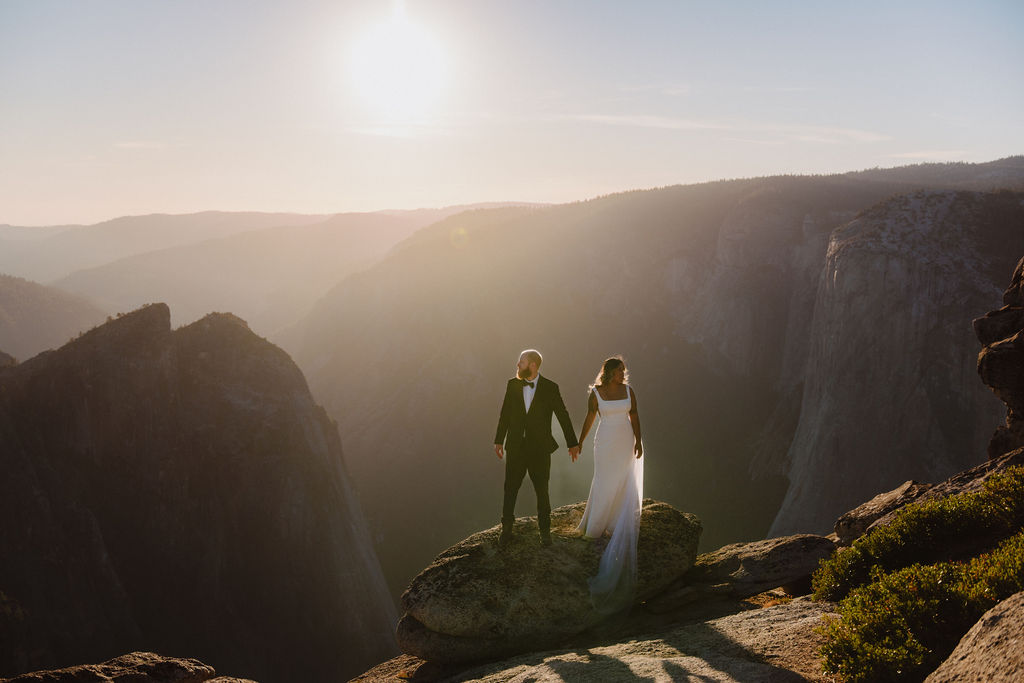 A couple in wedding attire stands on a rocky ledge, holding hands, with a mountainous landscape and bright sun in the background for their glacier point elopement