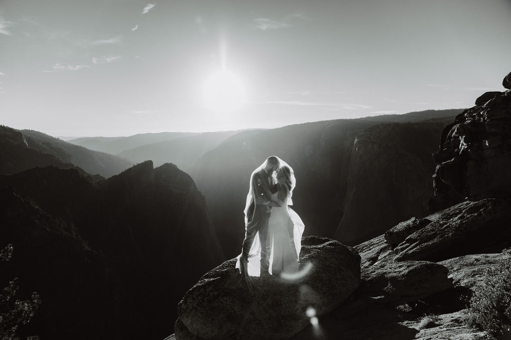 A couple in wedding attire stands on a rocky ledge, holding hands, with a mountainous landscape and bright sun in the background for their glacier point elopement