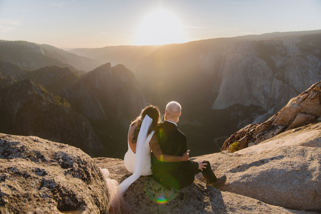 A bride and groom sit on a rocky ledge overlooking a mountainous landscape at sunset.