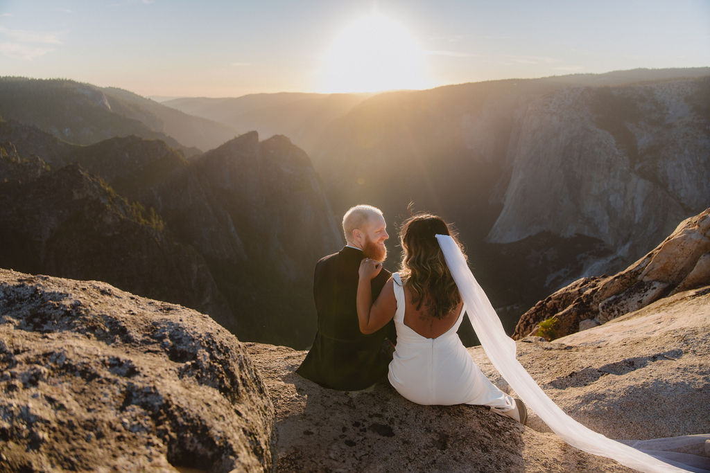 A bride and groom sit on a rocky ledge overlooking a mountainous landscape at sunset.