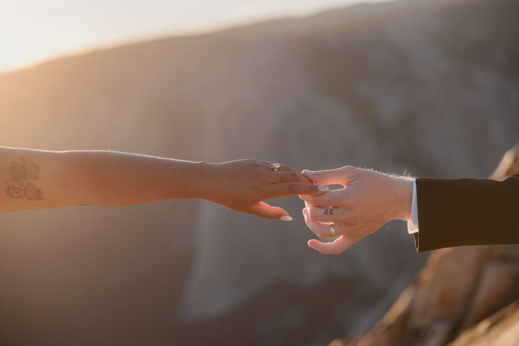 A bride and groom sit on a rocky ledge overlooking a mountainous landscape at sunset.