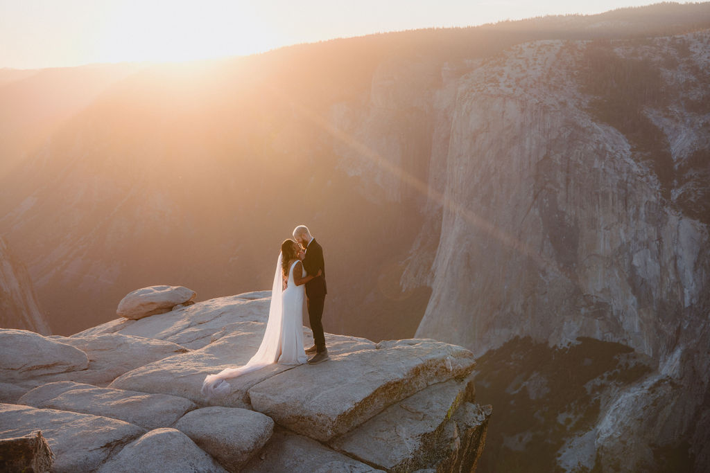 A couple in wedding attire stands on a rocky cliff at sunset, overlooking a vast mountain landscape for a glacier point elopement