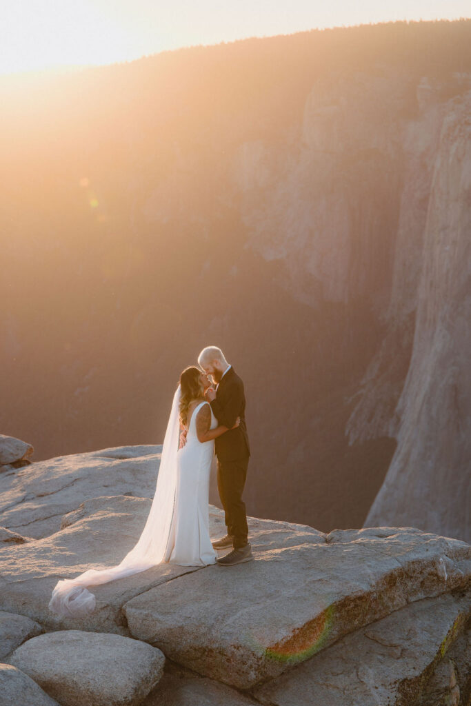 A bride and groom stand on a rocky cliff at sunset, wrapped in a veil, with mountains and a bright sun in the background for an elopement in Yosemite