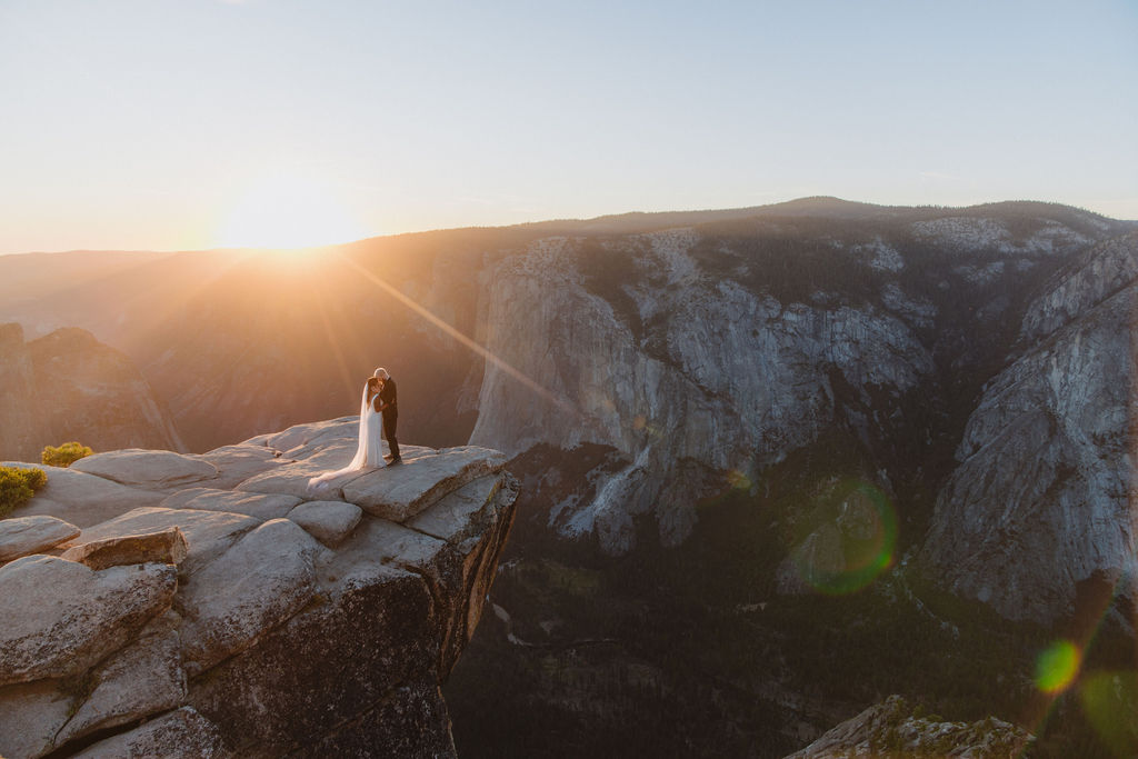 A couple in wedding attire stands on a rocky cliff at sunset, overlooking a vast mountain landscape for a glacier point elopement