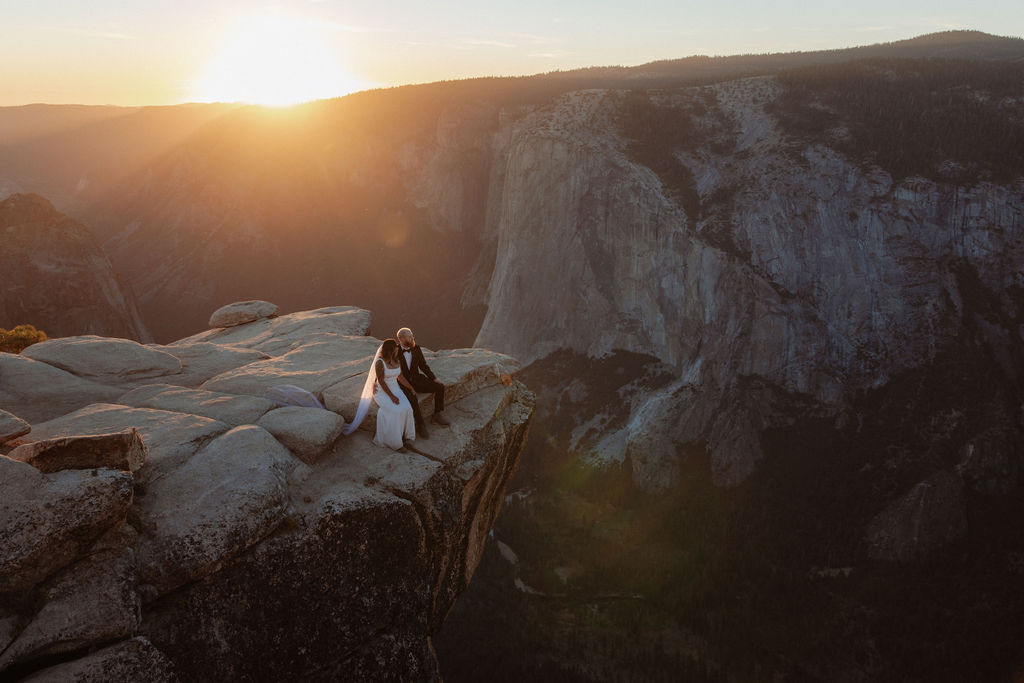 A couple stands on the edge of a rocky cliff at sunset, facing each other with distant mountains and a bright sun in the background for their glacier point elopement