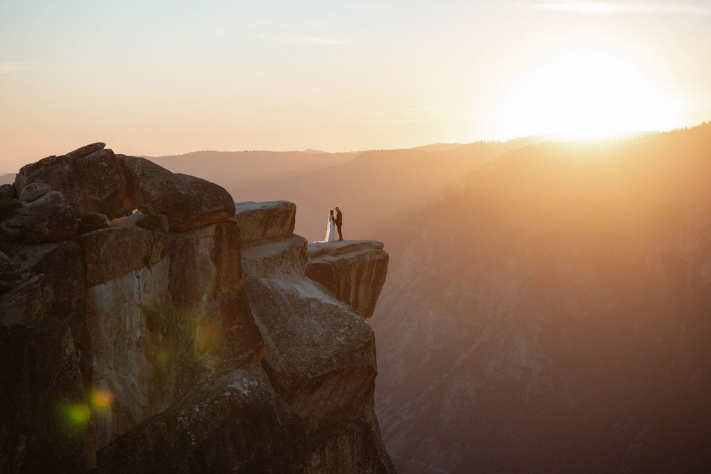 A couple stands on the edge of a rocky cliff at sunset, facing each other with distant mountains and a bright sun in the background for their glacier point elopement