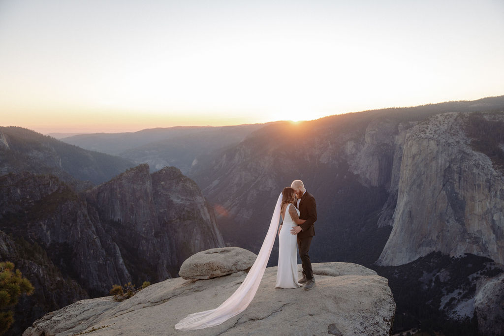 A bride and groom stand on a rocky cliff at sunset, wrapped in a veil, with mountains and a bright sun in the background for an elopement in Yosemite