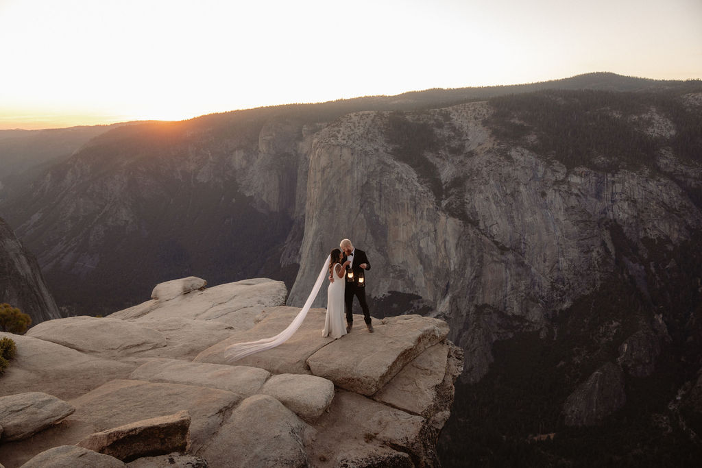 A bride and groom stand on a rocky cliff edge at sunset with mountains and a valley in the background for a glacier point elopement