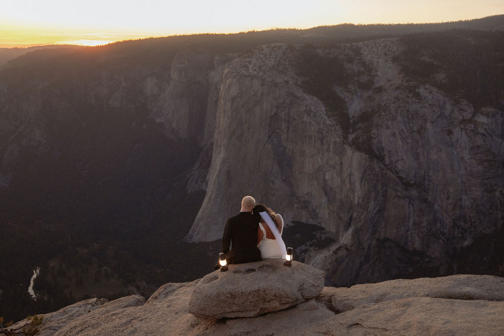 A bride and groom stand on a rocky cliff edge at sunset with mountains and a valley in the background for a glacier point elopement