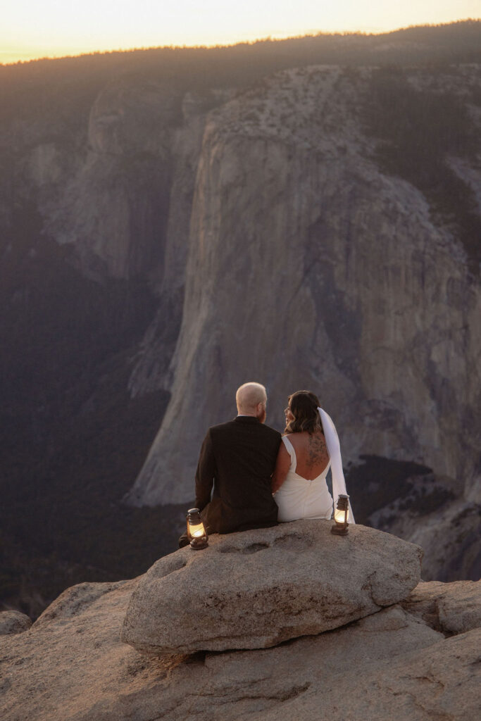 A couple in wedding attire sits on a rocky ledge at sunset, facing a vast mountain landscape, with lanterns placed beside them.