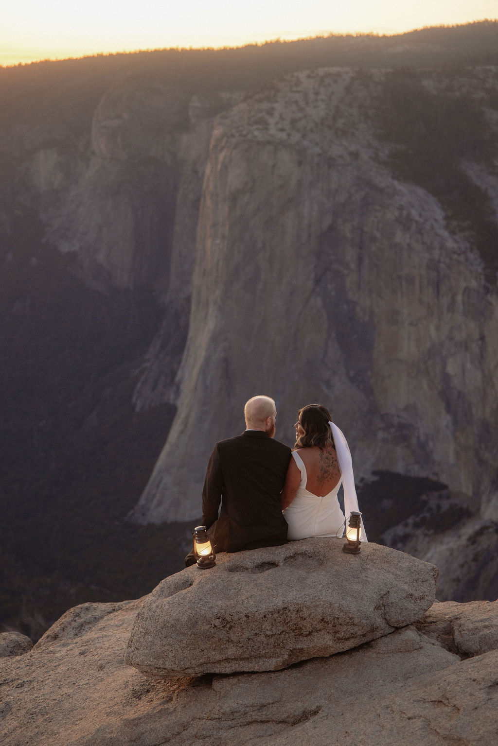 A couple in wedding attire sits on a rocky ledge at sunset, facing a vast mountain landscape, with lanterns placed beside them.