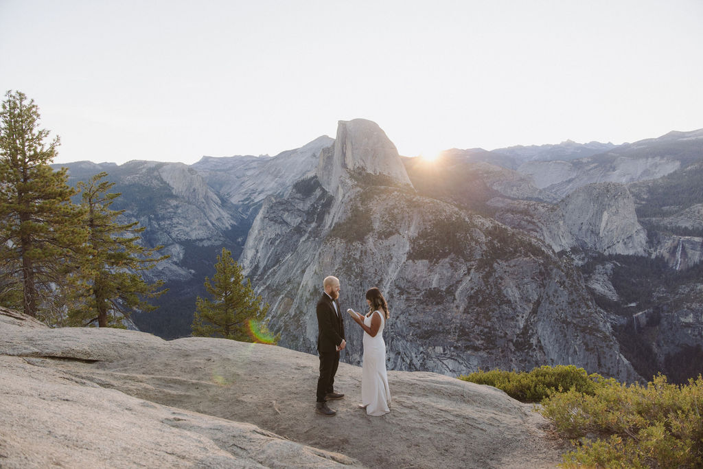 A couple stands on a rocky overlook exchanging vows at sunset, with mountains and Half Dome in the background.