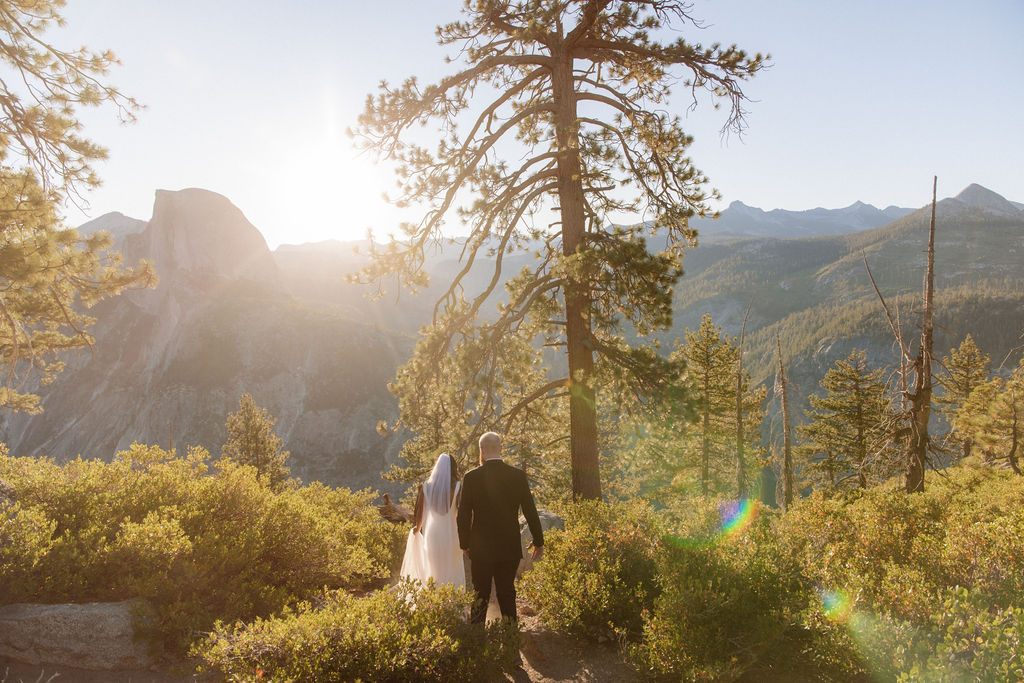 A couple in wedding attire kisses in a sunlit, mountainous landscape with trees and rocky cliffs in the background for a glacier point elopement