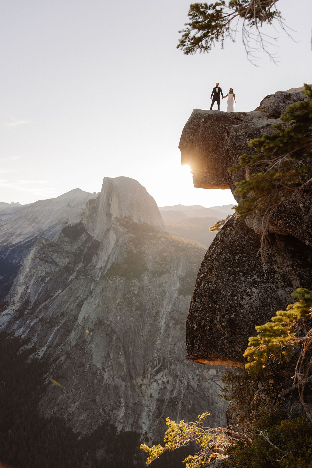 A couple in wedding attire kisses in a sunlit, mountainous landscape with trees and rocky cliffs in the background for a glacier point elopement