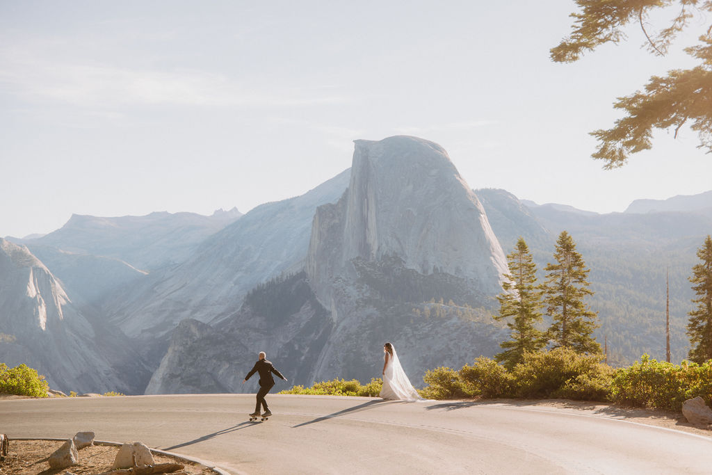 A couple in wedding attire walks hand in hand down a paved road lined with trees, with a large mountain visible in the background.