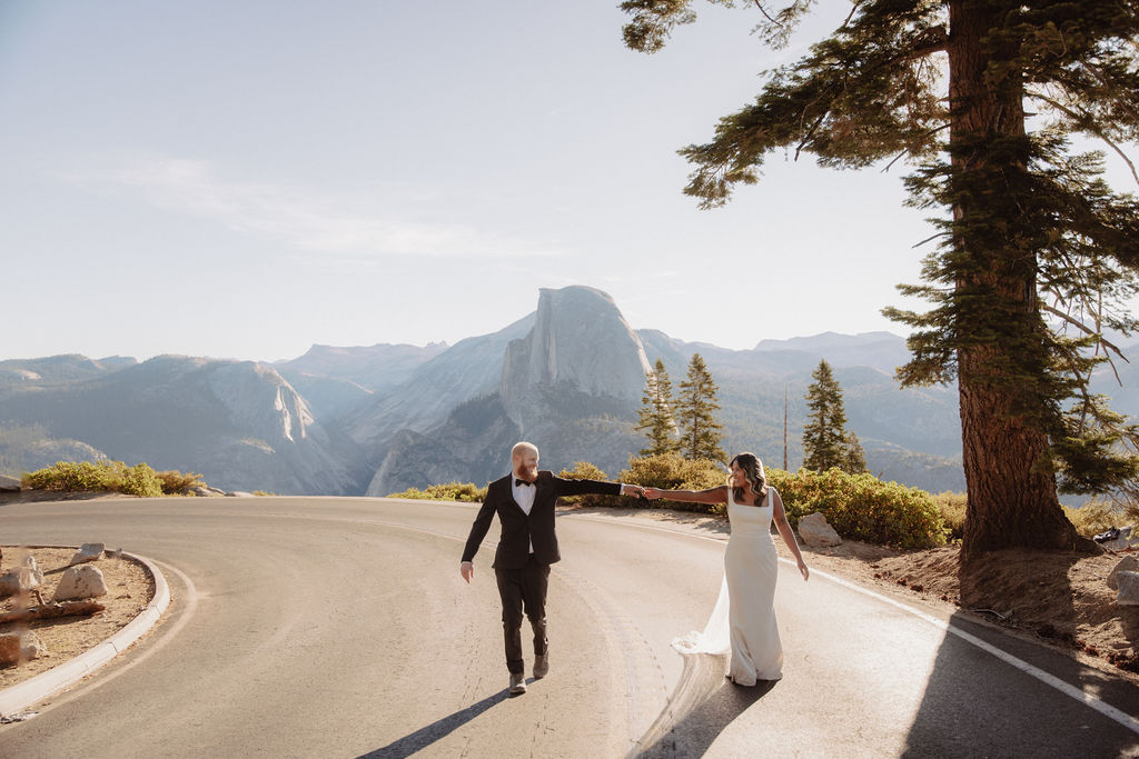 A couple in wedding attire walks hand in hand down a paved road lined with trees, with a large mountain visible in the background.