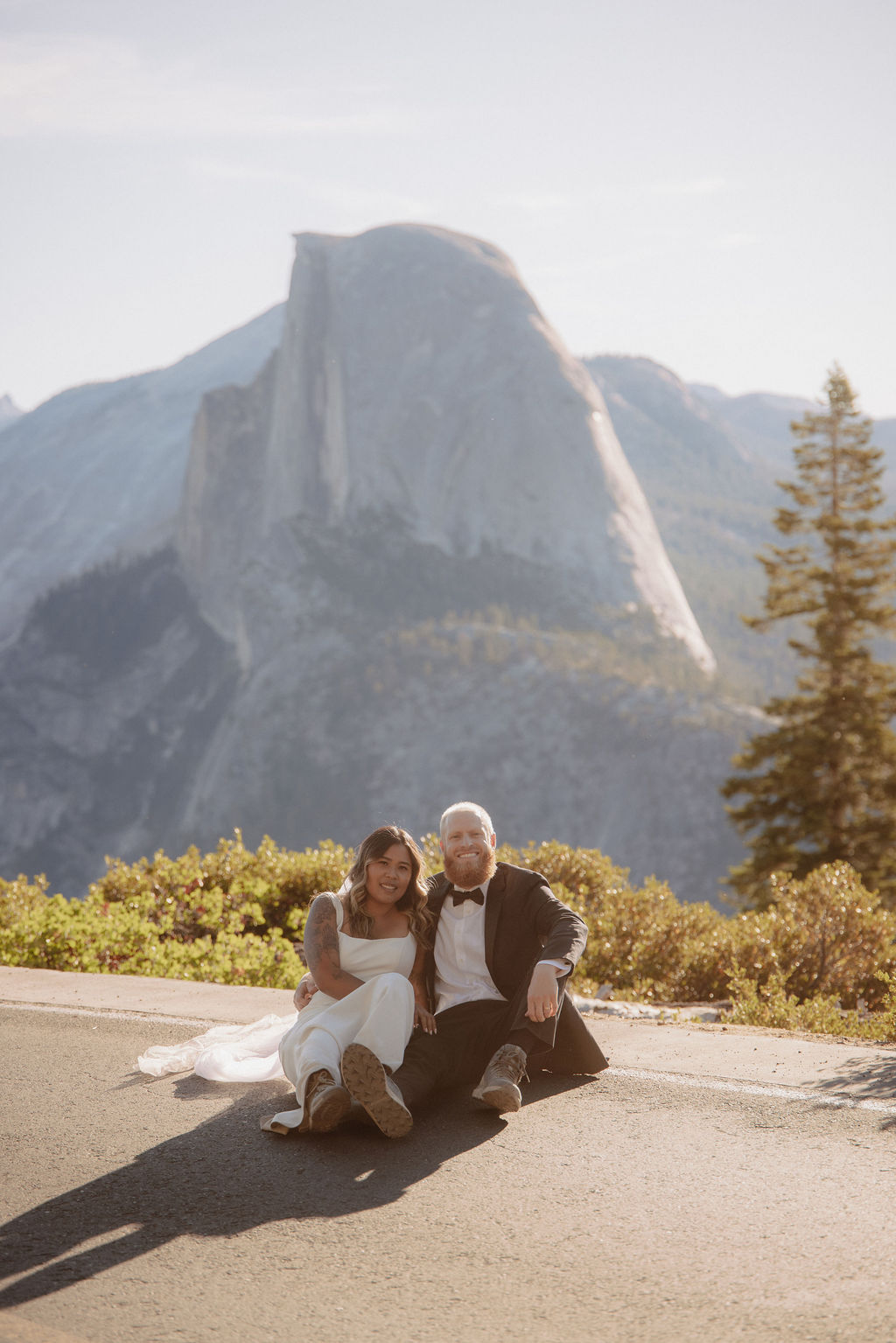 A couple in wedding attire kisses in a sunlit, mountainous landscape with trees and rocky cliffs in the background for a glacier point elopement