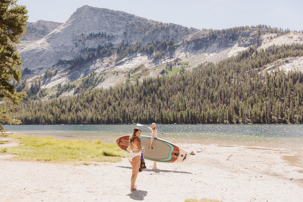 A man and woman sit together on a paddleboard in a clear lake, with forested mountains in the background under a clear sky.