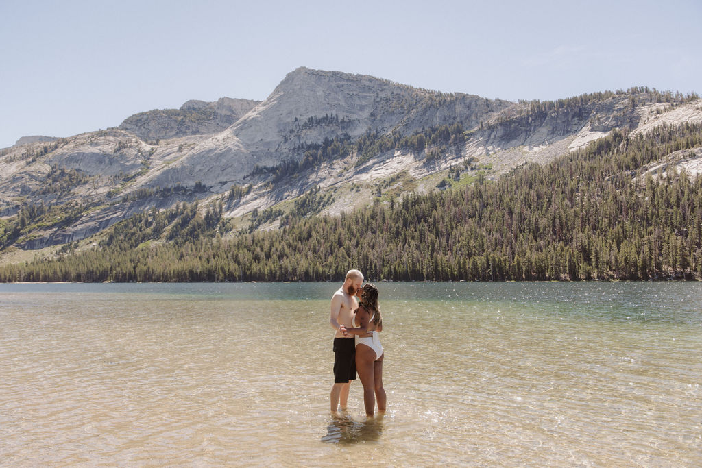 A man and woman sit together on a paddleboard in a clear lake, with forested mountains in the background under a clear sky.