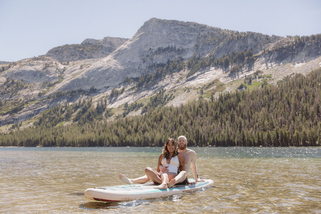 A man and woman sit together on a paddleboard in a clear lake, with forested mountains in the background under a clear sky.