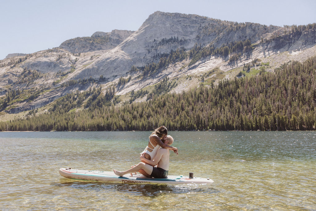A man and woman sit together on a paddleboard in a clear lake, with forested mountains in the background under a clear sky.