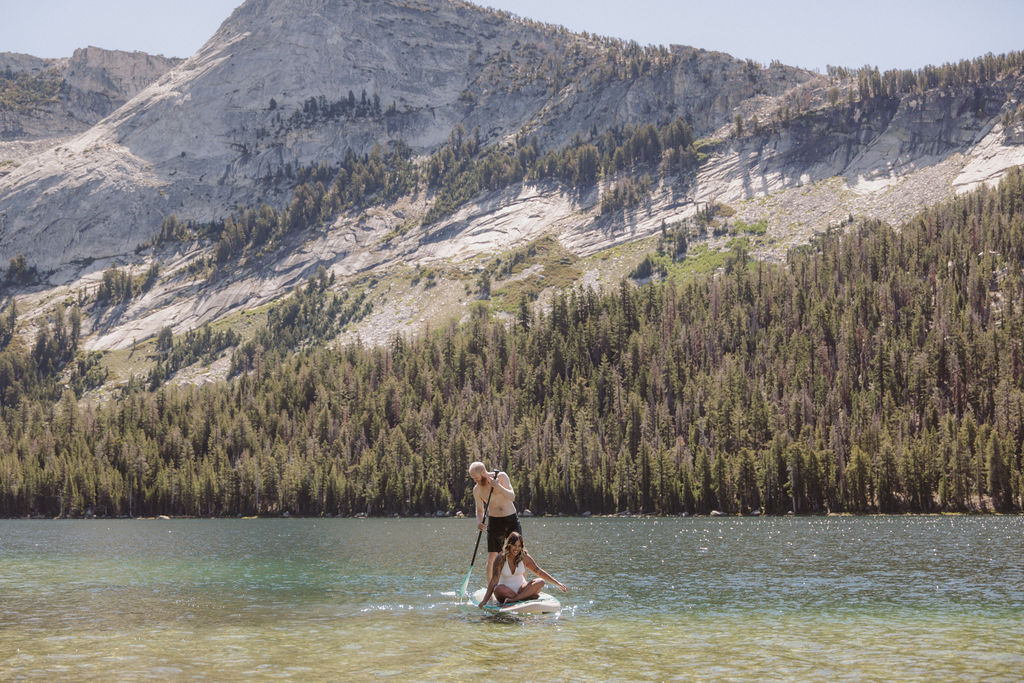 A man and woman sit together on a paddleboard in a clear lake, with forested mountains in the background under a clear sky.