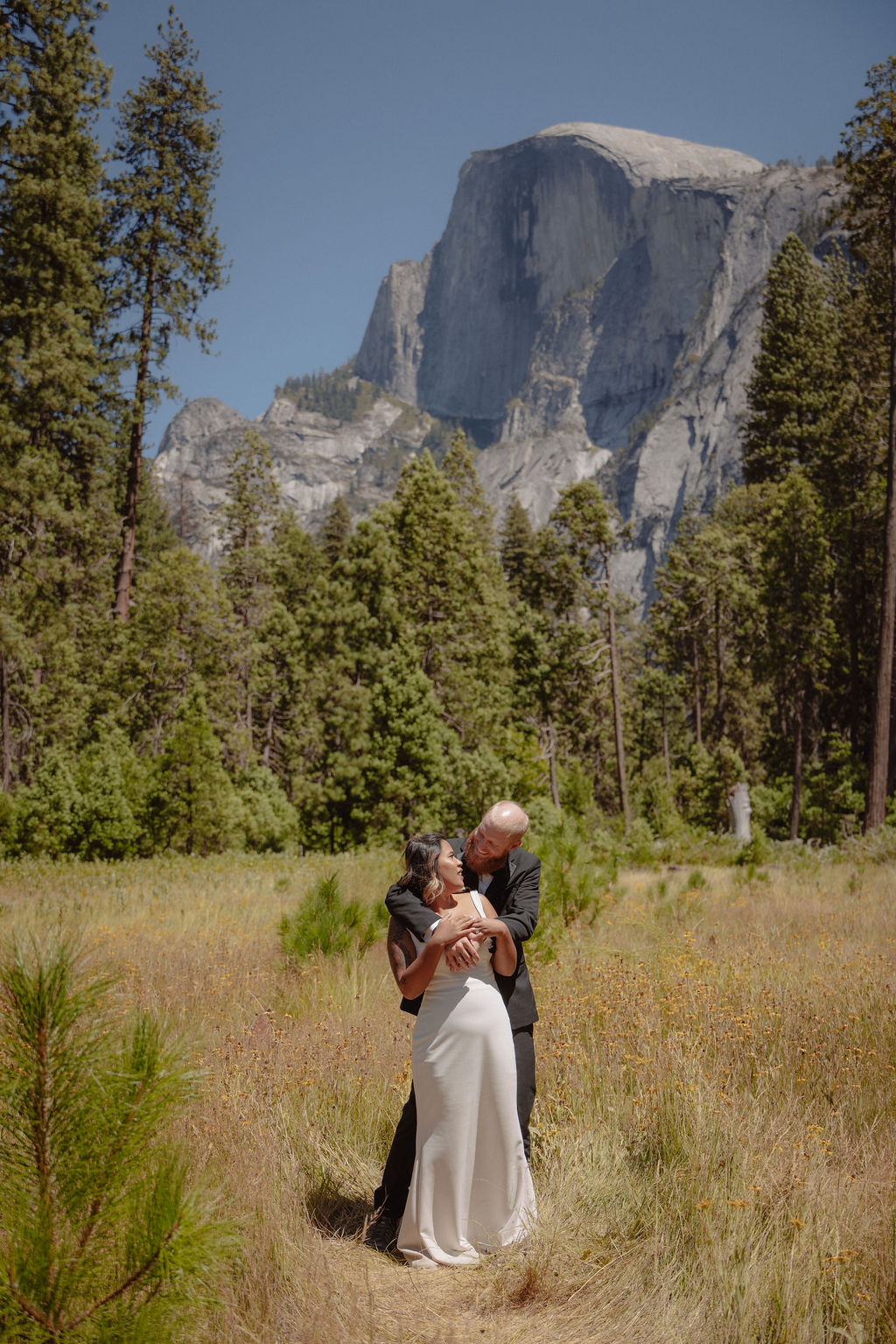 A man in a suit and a woman in a white dress stand in a meadow holding newspapers in front of their faces, with a large rocky mountain in the background.