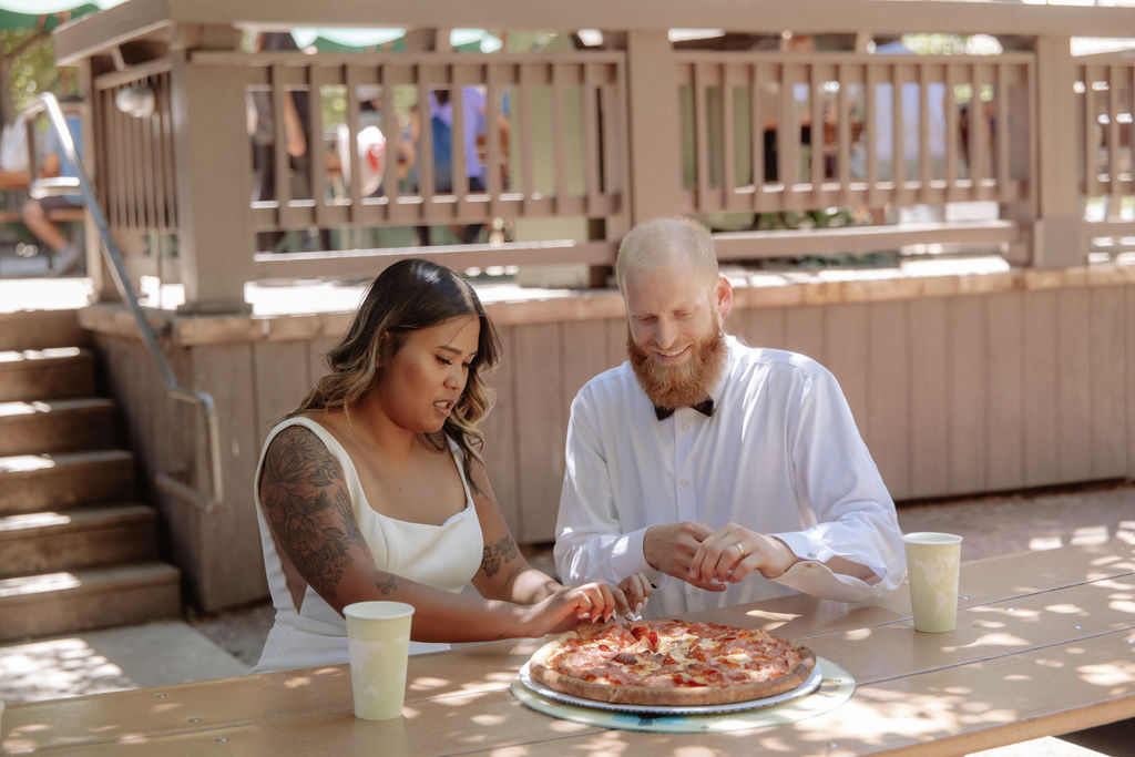 A woman and a man sit at an outdoor table sharing a pizza, each reaching for a slice. Two large drinks are on the table in front of them for a glacier point elopement 