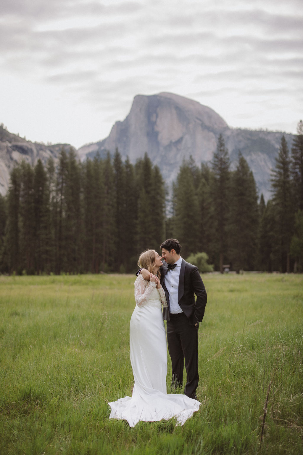 A bride and groom stand together in a grassy field with pine trees and a large mountain in the background, both facing each other and holding hands for their elopement in Yosemite | elopement budget