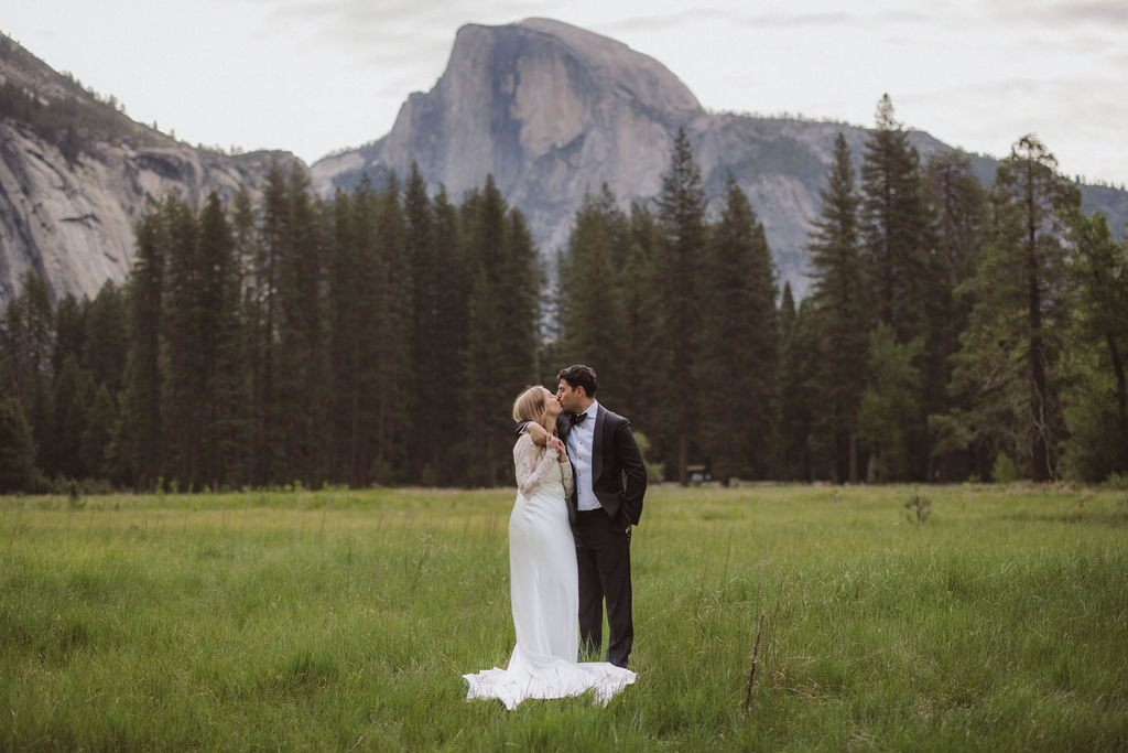 A bride and groom stand together in a grassy field with pine trees and a large mountain in the background, both facing each other and holding hands for their elopement in Yosemite | elopement budget