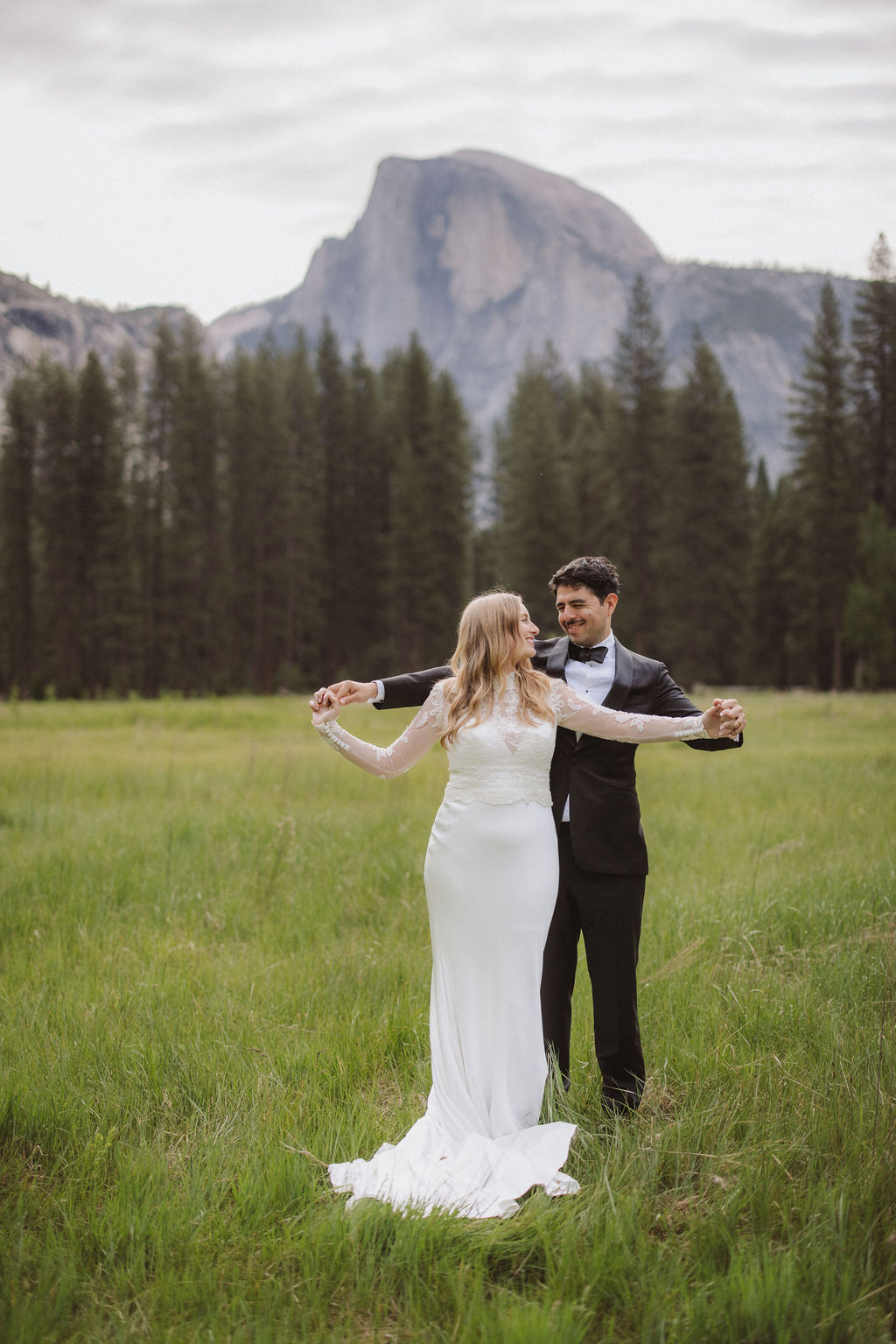 A bride and groom stand together in a grassy field with pine trees and a large mountain in the background, both facing each other and holding hands for their elopement in Yosemite | elopement budget
