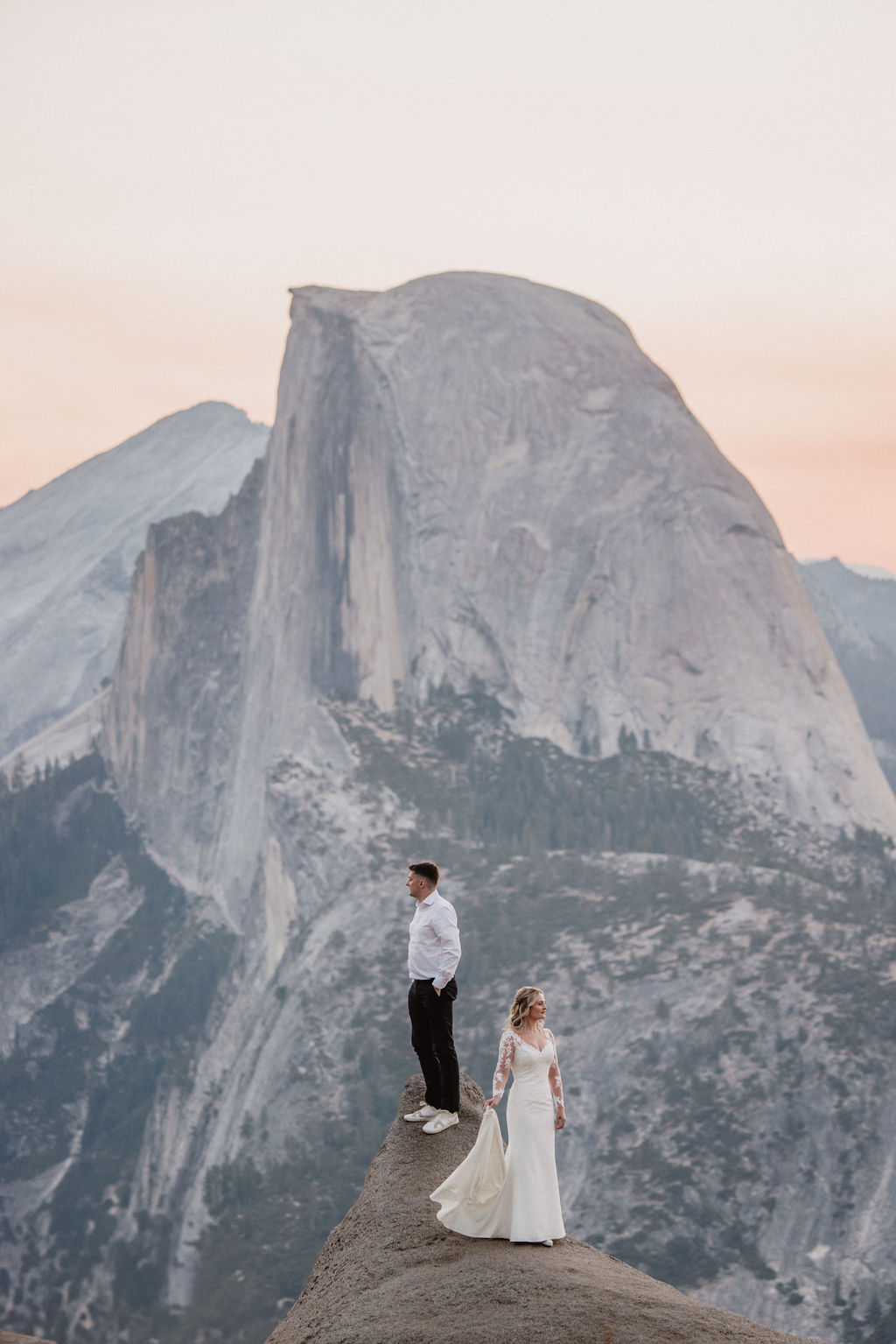 A couple dressed in formal attire stands on a cliff edge with Half Dome mountain in the background at sunset.